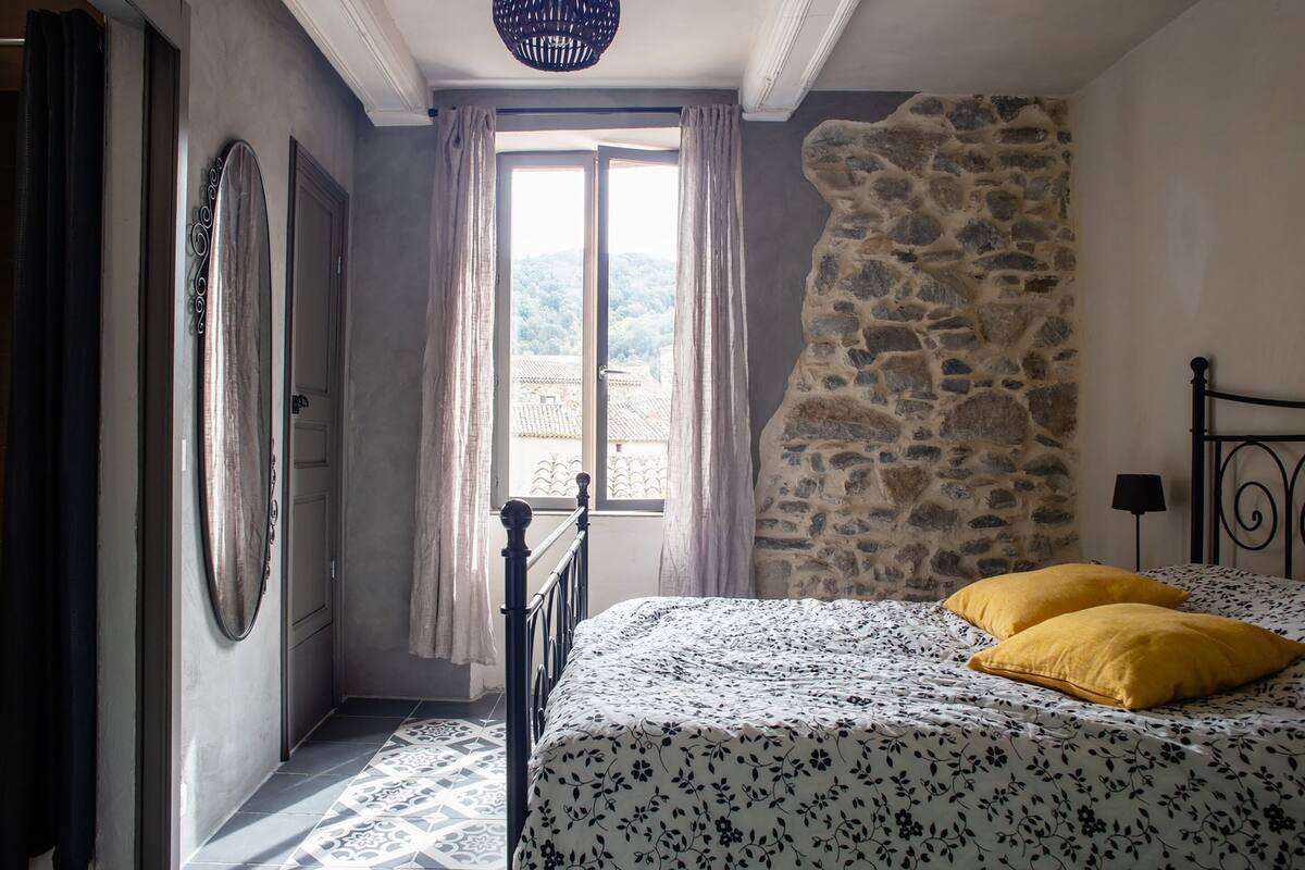 Un dormitorio con cama, pared de piedra, ventana con cortinas y vista a un pueblo.