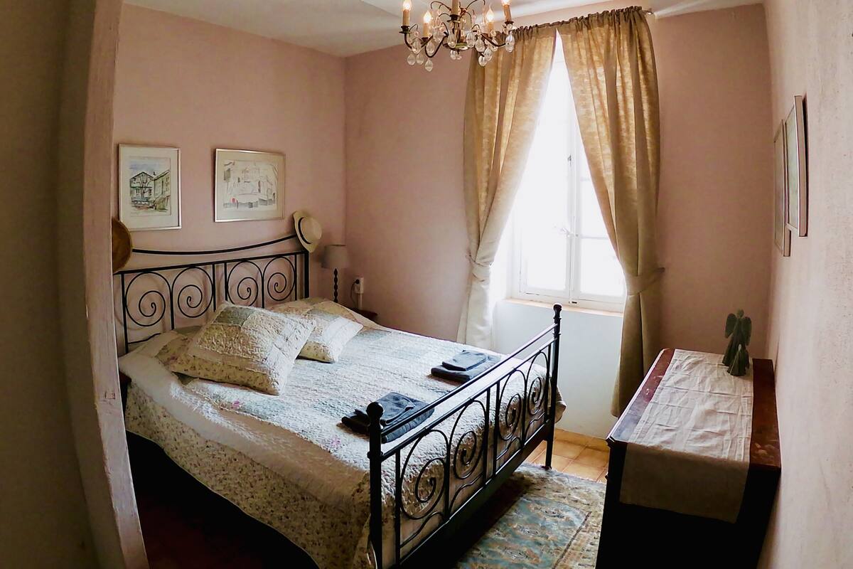 Un dormitorio con cama, mesita de noche, lámpara de araña y ventana con cortinas.