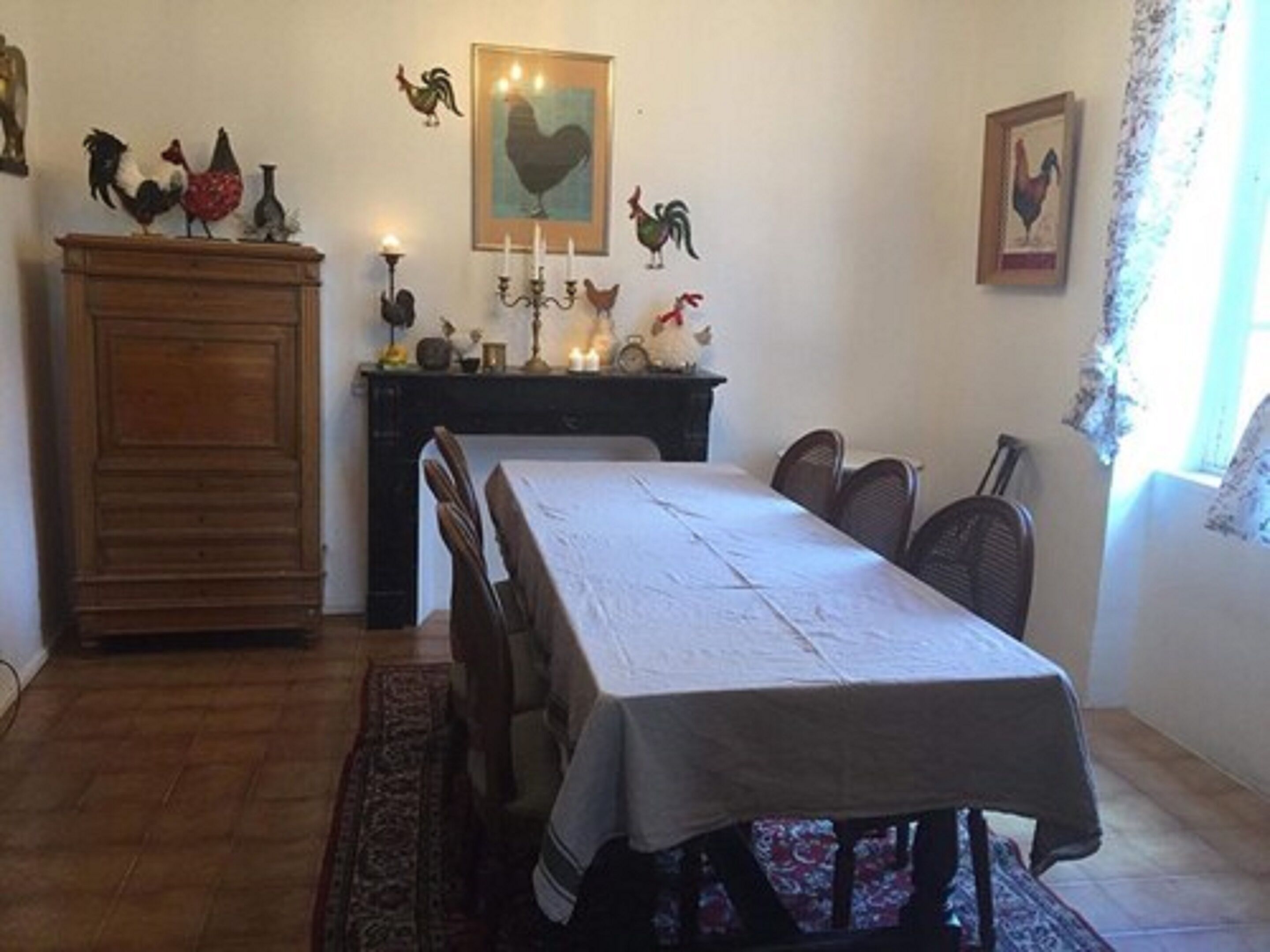 A dining room with a long table, chairs, a wooden cabinet, and wall-mounted decorations including roosters.