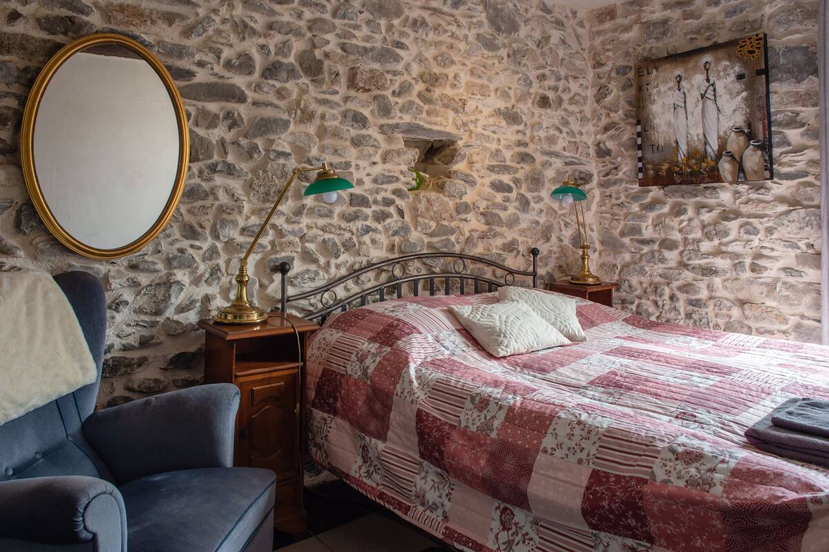 Un dormitorio con pared de piedra, una cama con colcha rojo y blanco, un sillón azul, una mesita de noche de madera con dos lámparas y un espejo.
