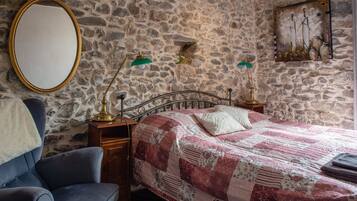 Een slaapkamer met een stenen muur, een bed met een rood-wit quilt, een blauwe fauteuil, een houten nachtkastje met twee lampen en een spiegel.