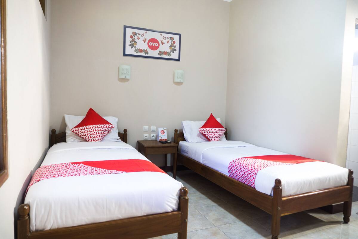 Two single beds with wooden frames, white bedding, and red and white patterned pillows. There is a framed sign with the word "OYO" on the wall, and two wall-mounted light switches.