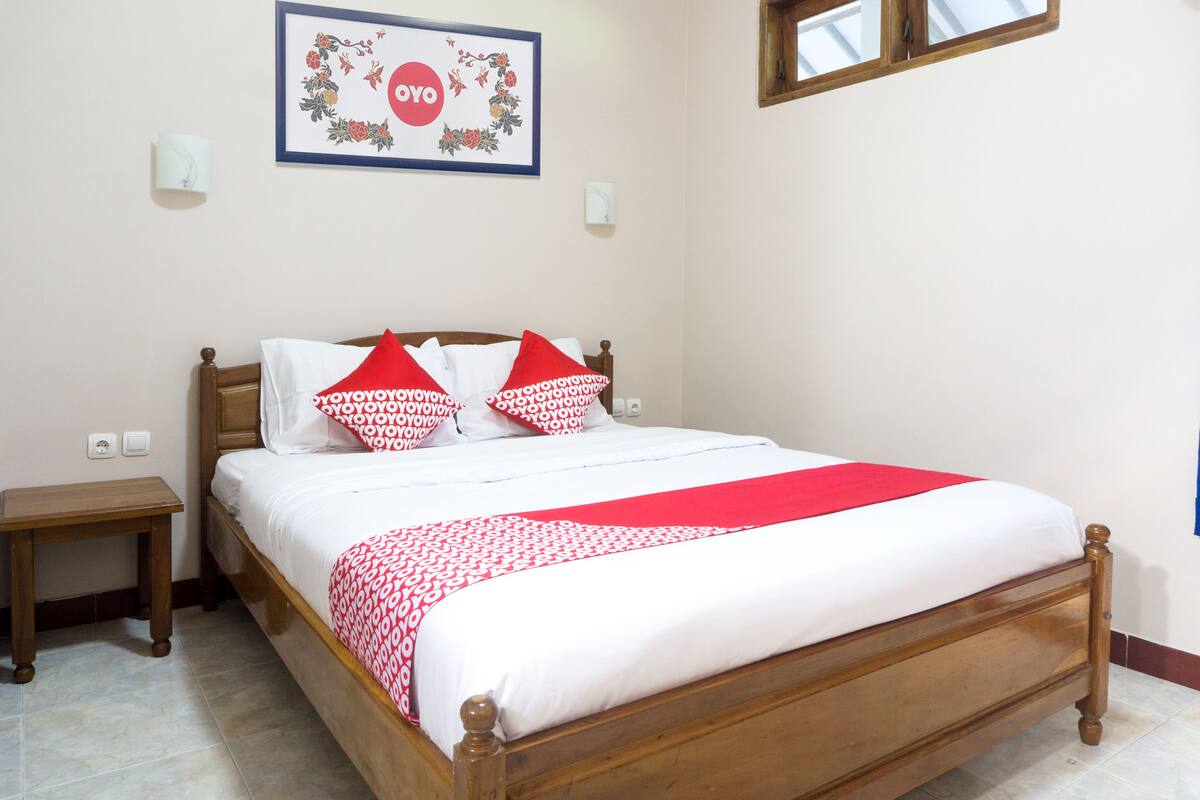 A neatly made bed with a red and white patterned bedspread and pillows, a wooden headboard, and a framed picture on the wall.