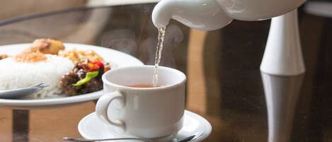 A teapot pouring tea into a cup on a glass table with a plate of food in the background.