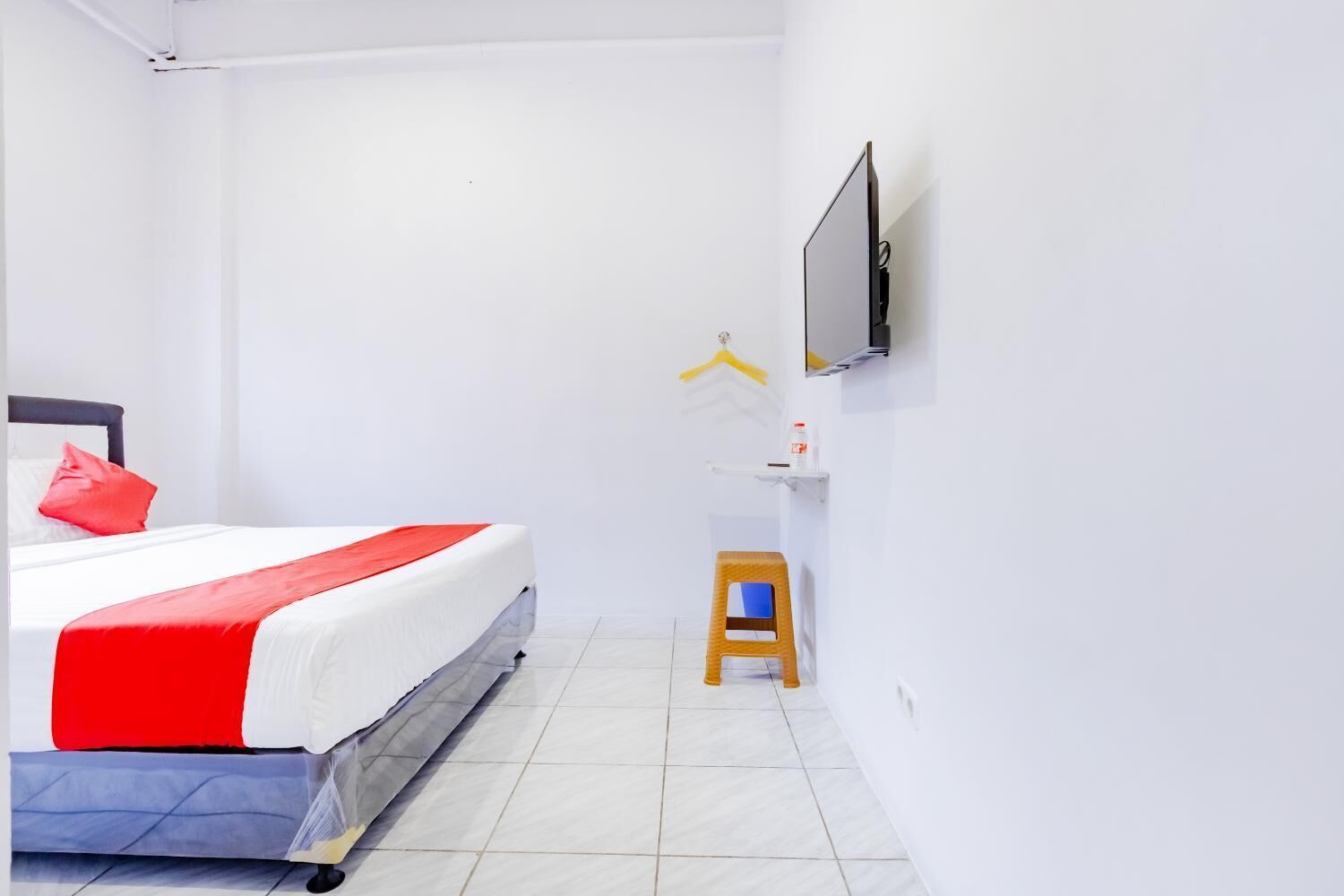 A hotel room with a bed, a small stool, a TV mounted on the wall, and a shelf with a bottle.