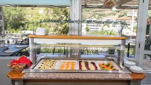 Buffet com diversos alimentos e vista para uma encosta através de amplas janelas.