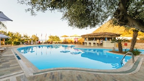 Piscina com espreguiçadeiras e guarda-sóis, rodeada por árvores e um edifício com telhado de palha.