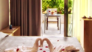 Quarto de hotel com uma cama adornada com arrumação de toalhas em formato de coração, uma mesinha com garrafa e copos, e vista para uma área externa com mesas e cadeiras através de uma porta de vidro.