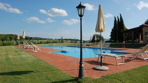 Piscina con sillas de descanso y sombrillas, rodeada de un patio de ladrillo y vegetación.