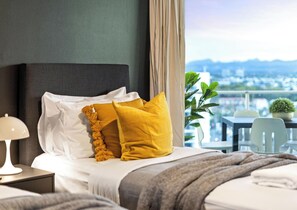 Habitación de hotel con una cama, una mesita de noche, una lámpara, vistas a un paisaje urbano a través de grandes ventanales y un balcón con sillas y una planta en maceta.