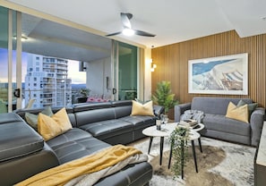 Una sala de estar moderna con un sofá de cuero negro, un sillón gris y una pequeña mesa de centro. Hay un ventanal con vistas a un paisaje urbano y un balcón.