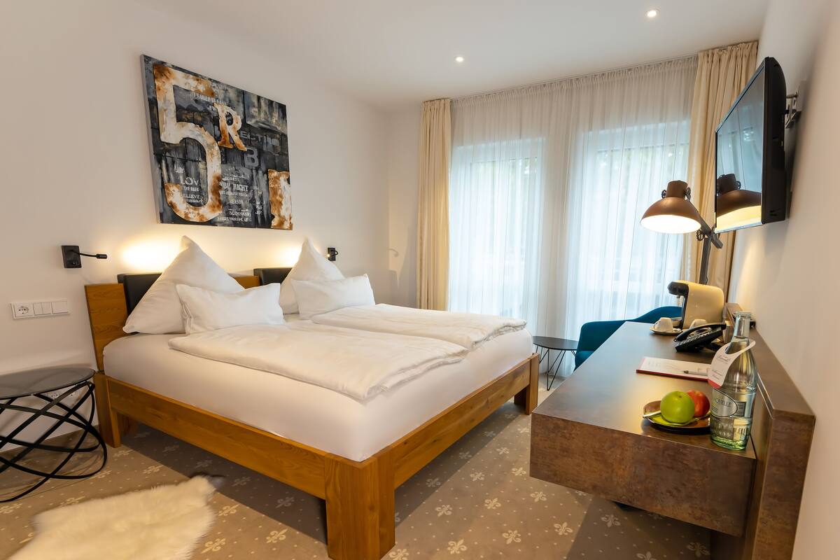 A modern hotel room with a large bed, bedside table, and a desk with a lamp.