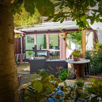 A covered outdoor seating area with wicker furniture, a table with a potted plant, and a view of the surrounding garden.