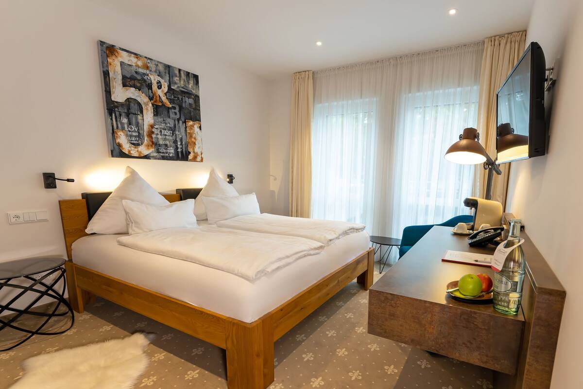A hotel room with a wooden bed, a desk with a lamp, a mirror, and a large window with curtains.