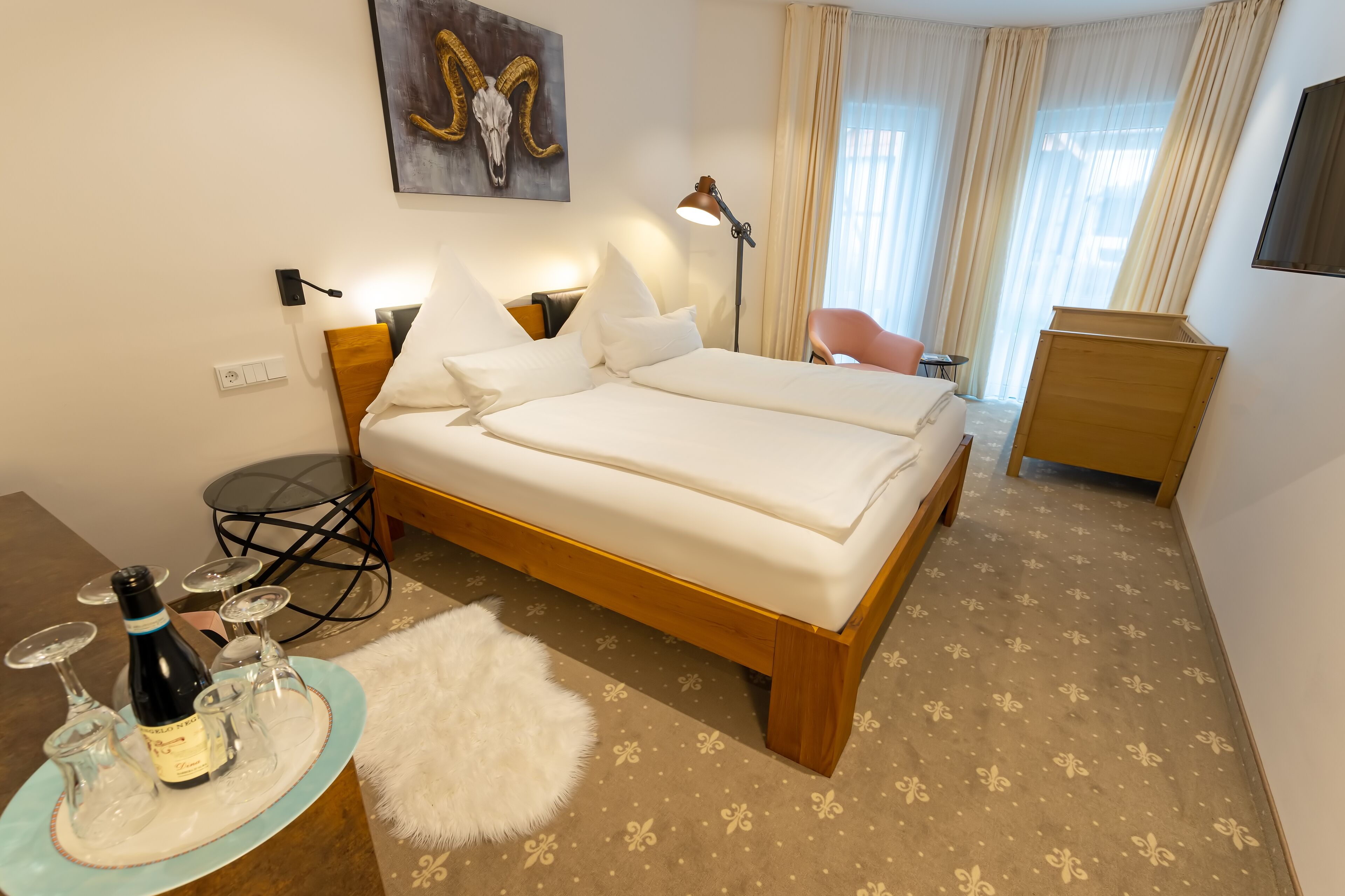 Ein Hotelzimmer mit Doppelbett, Schreibtisch mit Lampe, Stuhl, kleinem Tisch mit Flasche und Gläsern sowie einem Bild an der Wand.