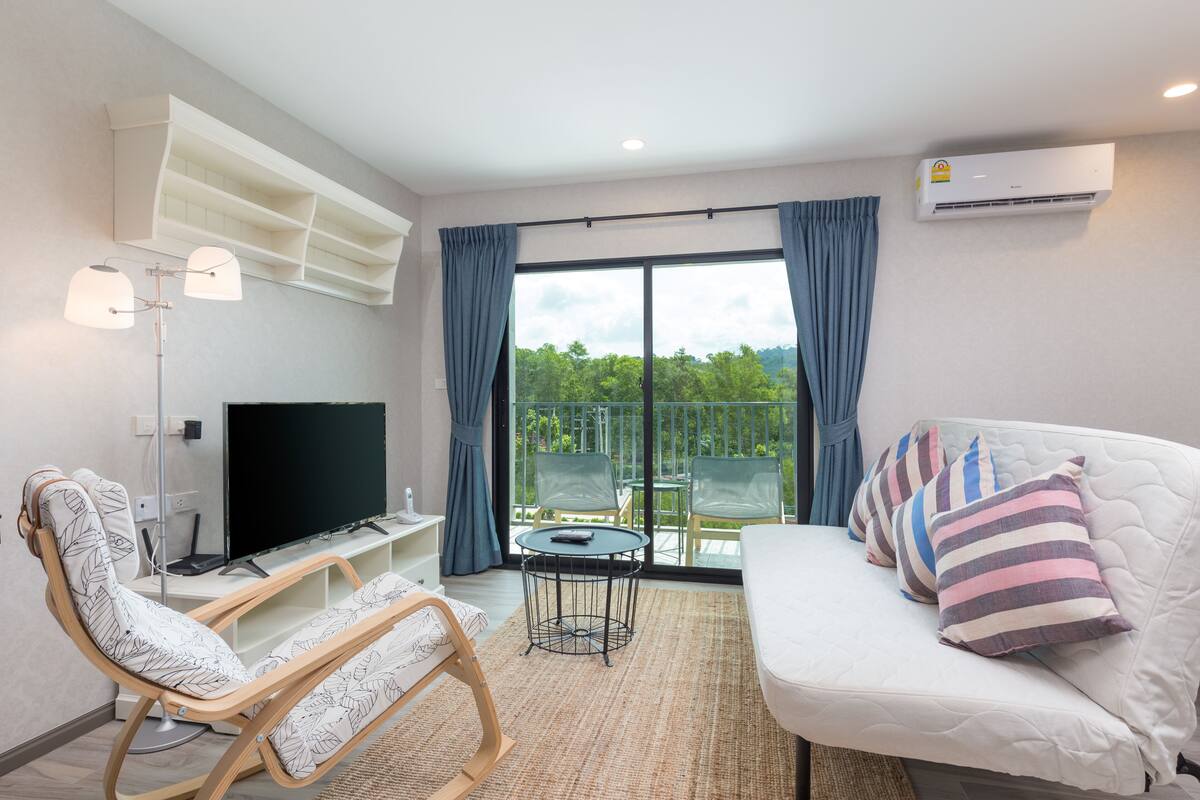 Un salon moderne comprenant un canapé, un fauteuil, un téléviseur à écran plat, une petite table et un balcon donnant sur un espace verdoyant.