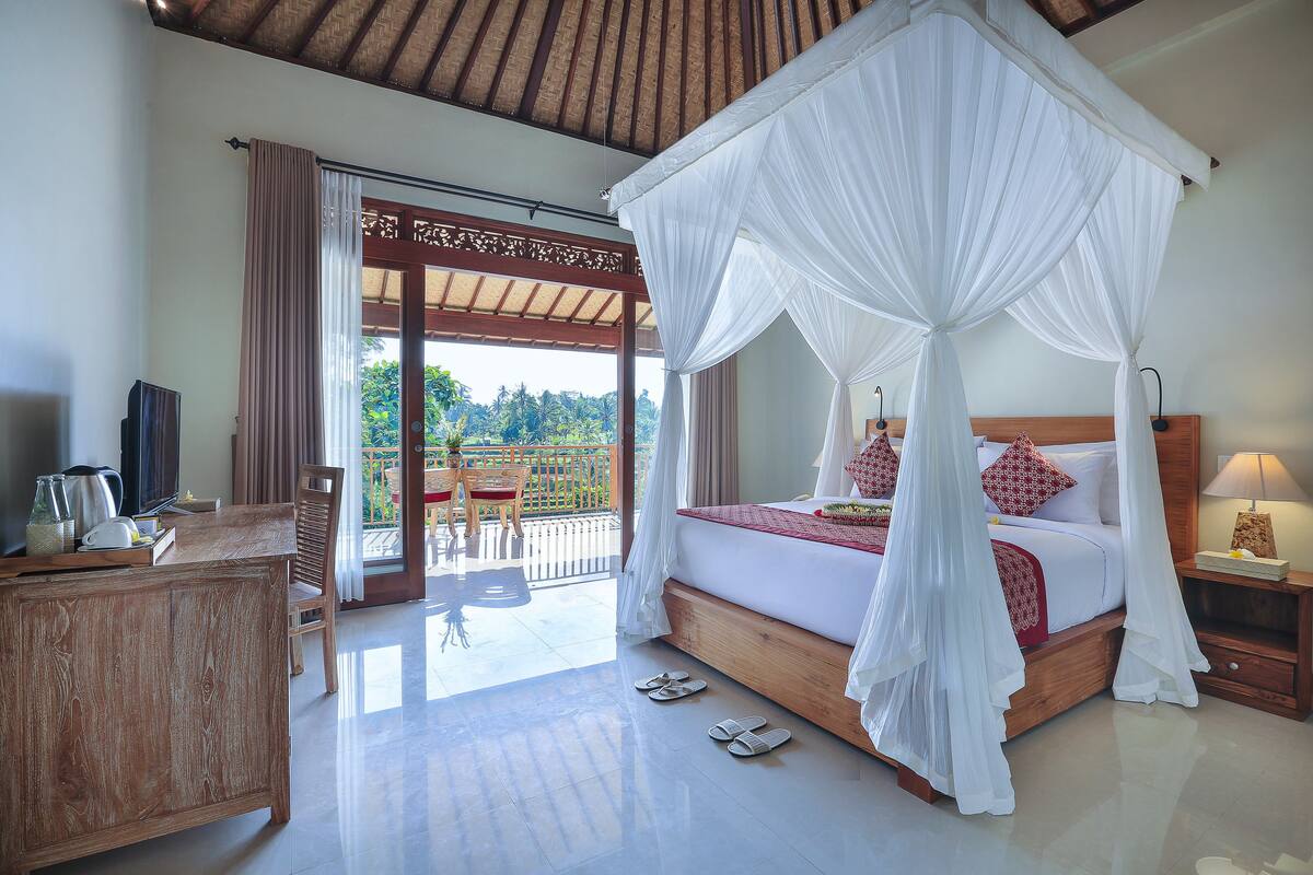 A bedroom with a four-poster bed, a wooden nightstand, a TV, and a balcony with a view of greenery.