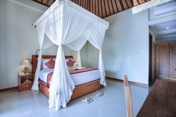 A bedroom with a four-poster canopy bed, a nightstand with a lamp, and slippers on the floor. - Nata Ubud (Ubud)