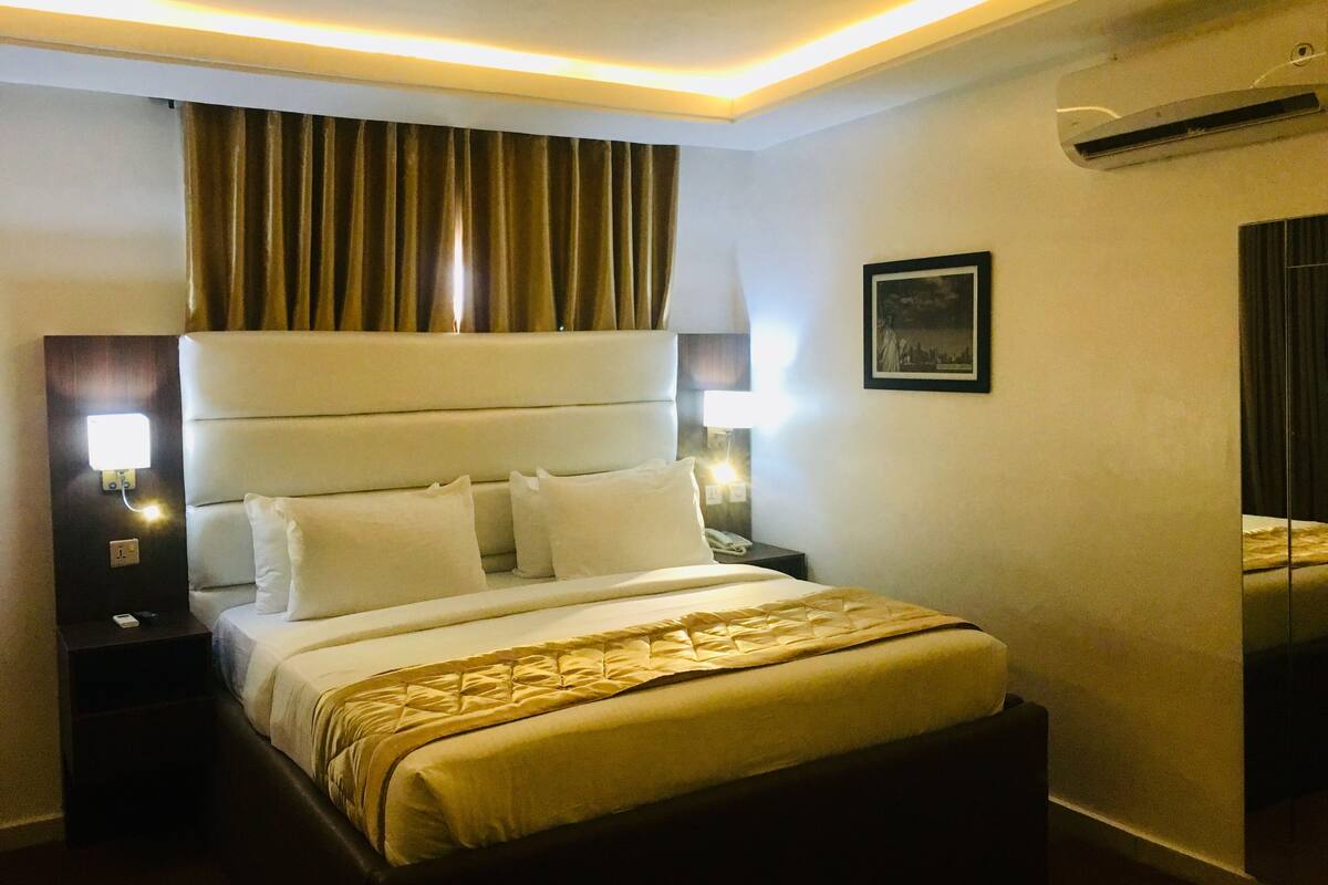 A hotel room with a large bed, two bedside tables, a nightstand, a wall-mounted air conditioner, and a framed picture on the wall.