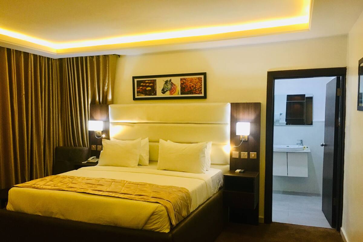 A hotel room with a large bed, bedside lamps, a nightstand, a headboard, a framed picture on the wall, and a bathroom visible through an open door.