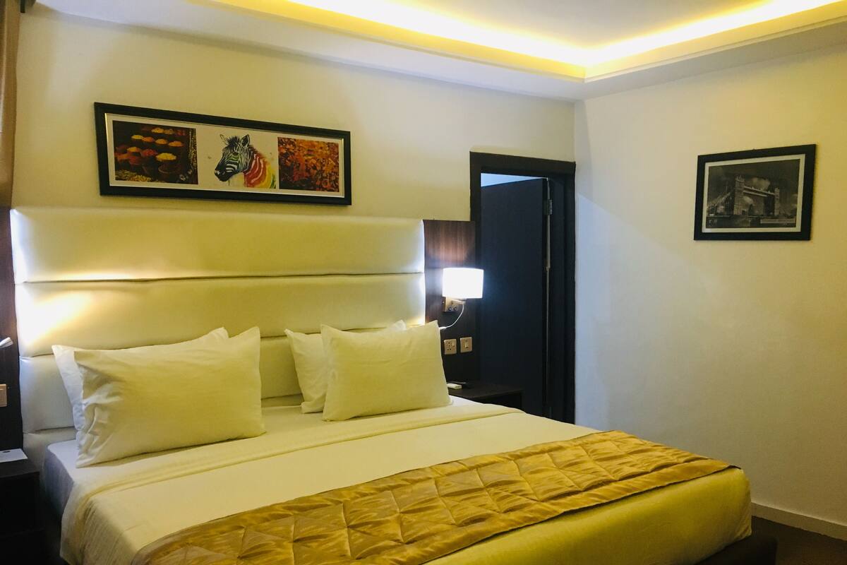 A hotel room with a large bed, a headboard, and a nightstand with a lamp. There are two framed pictures on the wall.