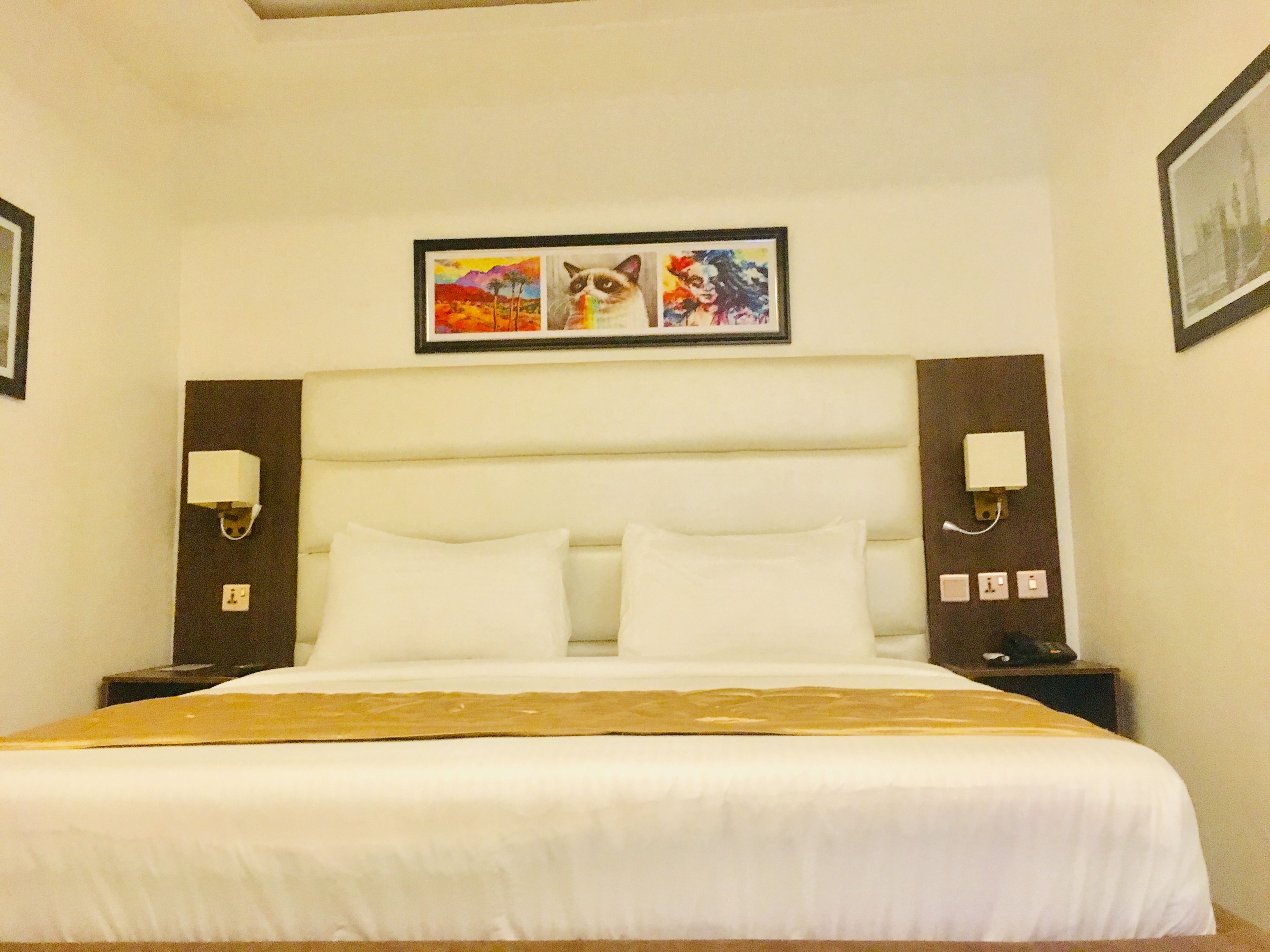 Ein Hotelzimmer mit einem großen Bett, weißer Bettwäsche, einem Kopfteil mit einem Gemälde, Nachttischlampen und einem Nachttisch mit Telefon und Lampe.