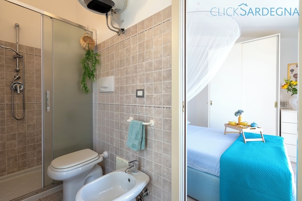 A bathroom with a shower, toilet, and bidet. There is a towel rack with a towel and a plant. The room has a bed with a blue blanket and a bedside table with cups and a vase.