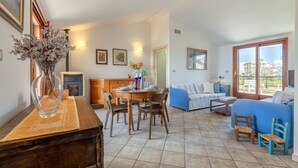 A spacious living area with a dining table set for four, a sofa, and a blue armchair. A fireplace is visible in the background, and there are framed pictures on the walls.