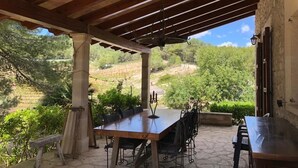 An outdoor patio with a wooden table and chairs, a stone floor, and a view of a hilly landscape.