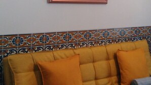 A yellow sofa with orange cushions and a blue blanket, a mirror on the wall, and patterned tiles below.