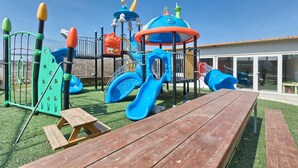 A playground with slides and climbing structures, surrounded by a wooden deck and a building with large windows. - Apartments Lovric - Apt 2 (Pula)