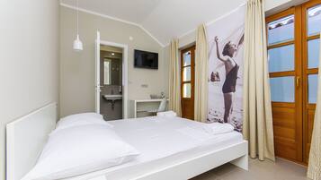 Un dormitorio con cama, televisor montado en la pared, escritorio y baño visible a través de una puerta abierta.