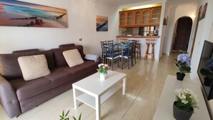 Una sala de estar con un sofá marrón, una mesa de centro blanca y un comedor con una cocina al fondo.