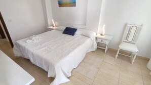 Una cama bien hecha con un cojín azul, dos mesitas de noche con lámparas, una silla blanca y un cuadro en la pared.
