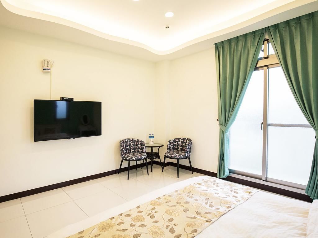 A hotel room with a flat-screen TV mounted on the wall, two patterned chairs around a small table, a large bed with a floral-patterned rug, and a sliding glass door with green curtains.