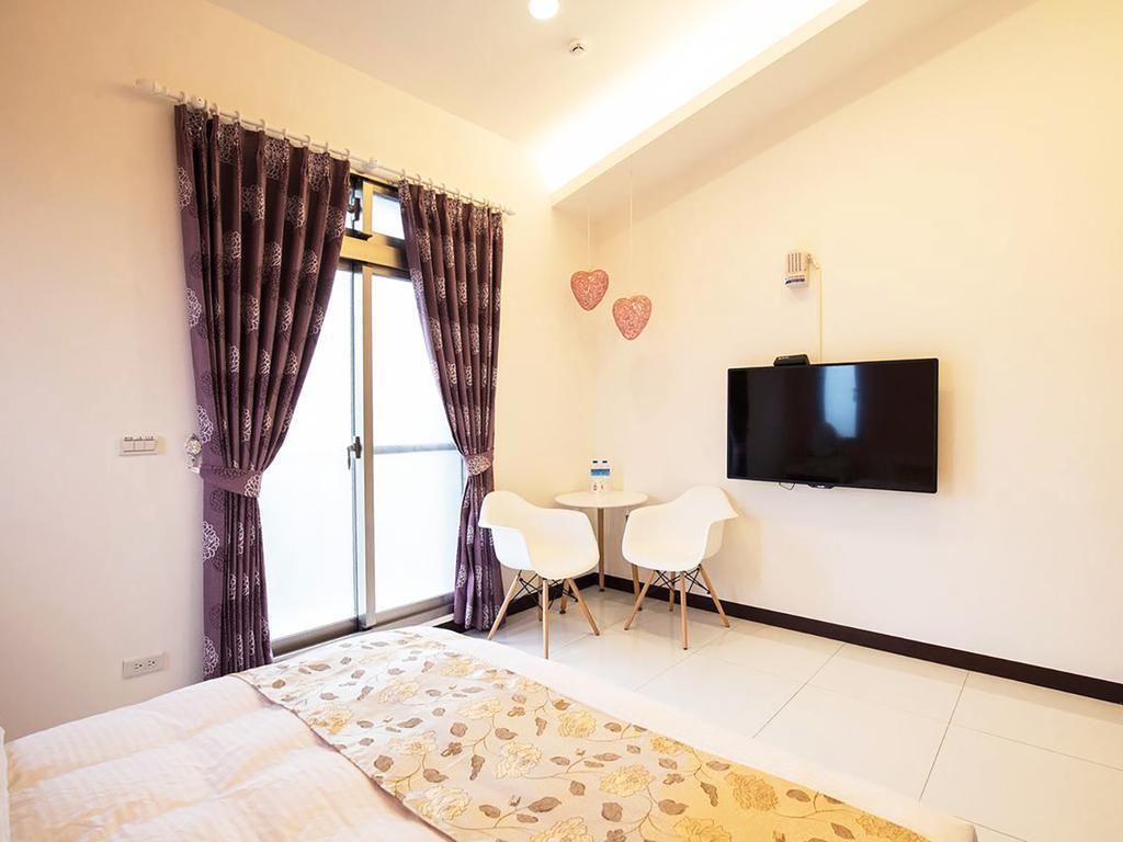 A hotel room with a bed, a TV mounted on the wall, two white chairs, a small table, and a window with curtains.