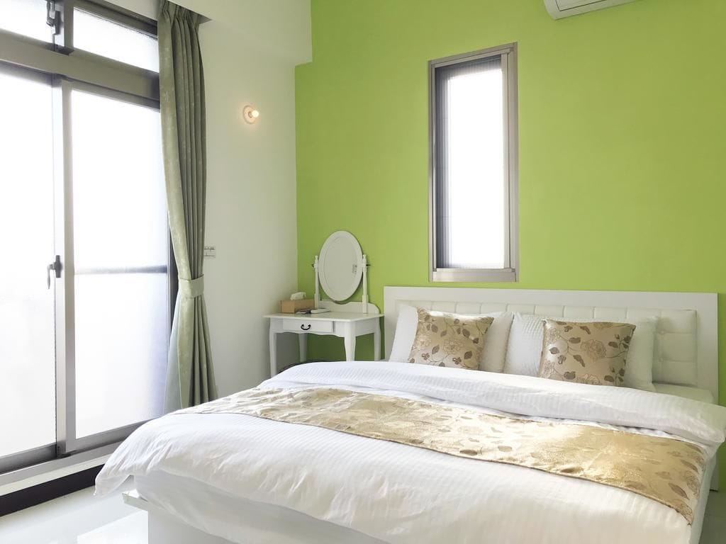A bedroom with a bed, a bedside table with a mirror, a window, and a view of the outside.