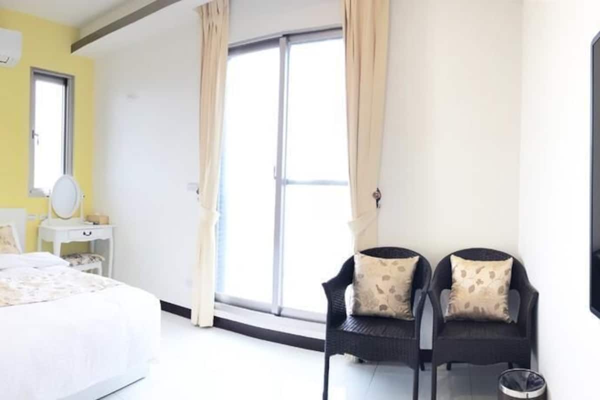 Een hotelkamer met een bed, nachtkastje, stoel en een groot raam.
