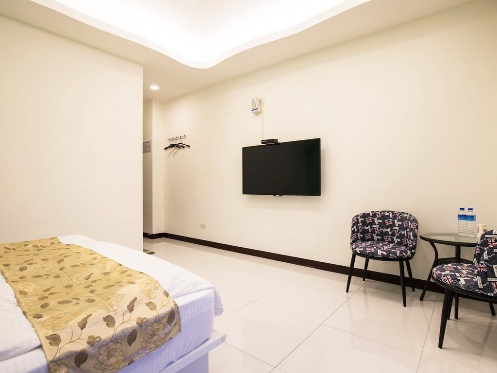 A hotel room with a bed, a TV mounted on the wall, a chair, and a small table with a water bottle.