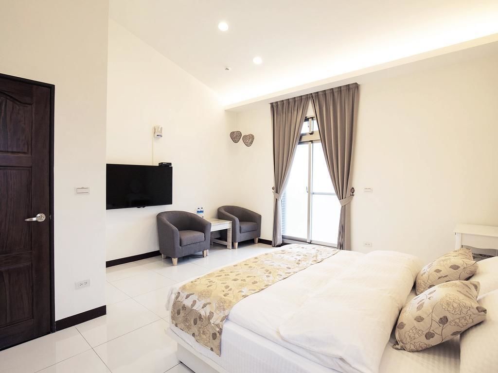 A modern hotel room with a large bed, a desk, two chairs, a flat-screen TV, and a window with curtains.