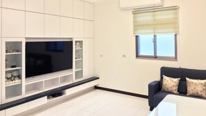 En moderne stue med et fladskærms-tv, indbyggede reoler, en sort sofa og et vindue med persienner.