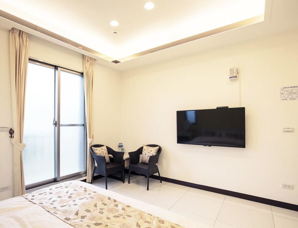 A hotel room with a bed, a TV mounted on the wall, a small table with chairs, and a sliding glass door leading to a balcony.