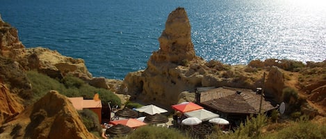 Une zone côtière caractérisée par un terrain rocheux et un petit bâtiment doté d’un toit en tuiles et de plusieurs parasols.