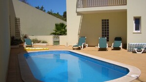 En swimmingpool med en buet kant, omgivet af en fliseterrasse. Der er liggestole og et lille bord i nærheden af poolen.