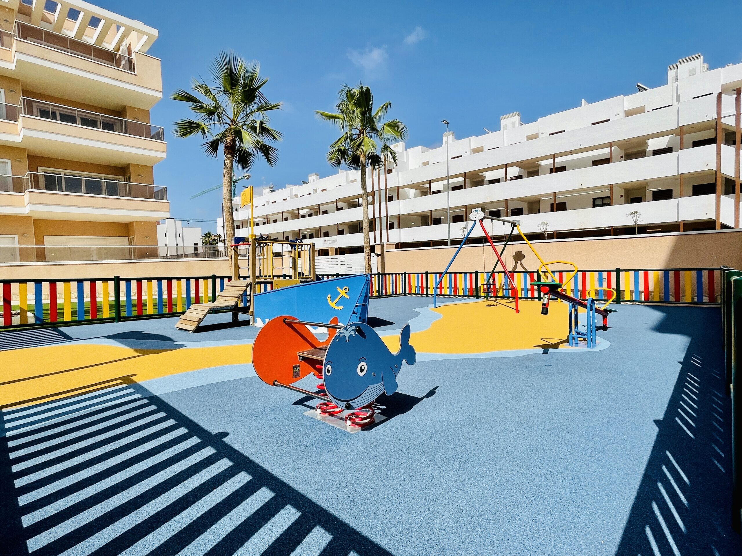 Un terrain de jeux avec des équipements colorés et des palmiers, le tout sur fond de bâtiments modernes.