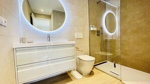 A modern bathroom with a glass shower enclosure, a white vanity with a round mirror, and a toilet. - Apartamento Azul (Villamartin) (Orihuela Costa)