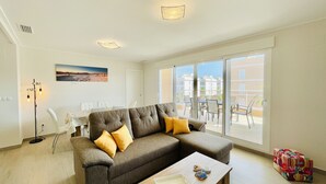A modern living room with a grey sectional sofa, a wooden coffee table, and a dining area with white chairs and a table.
