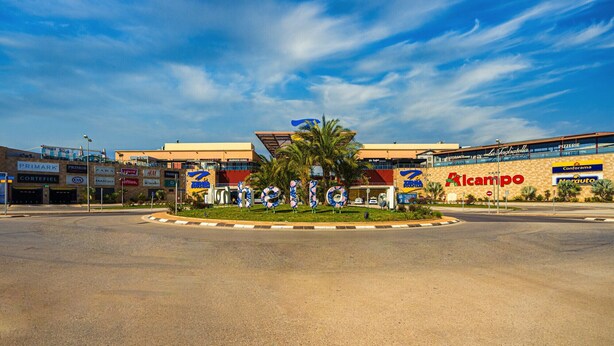 A commercial area with a central roundabout, surrounded by various businesses including a pharmacy, a fast-food restaurant, and a pizzeria. - Apartamento Azul (Villamartin) (Orihuela Costa)