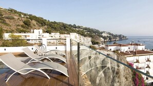 Une terrasse sur le toit, dotée de chaises longues, d’une plateforme en bois et offrant une vue sur les bâtiments côtiers et la mer.