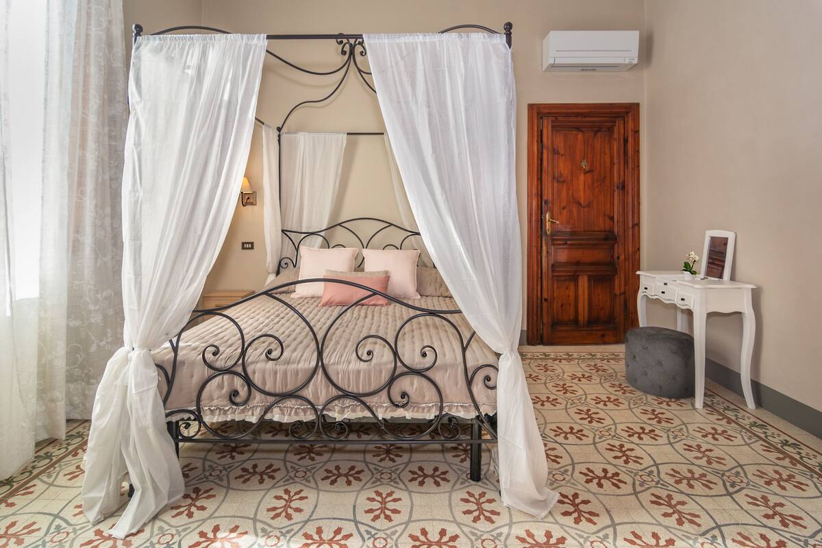 A four-poster bed with white curtains, a wooden door, a small white table, and patterned floor tiles.